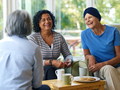 Two women laughing together while chatting with another person over tea and biscuits in a bright living room